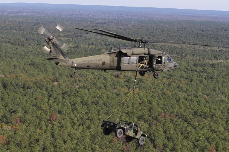 A helicopter carries a military vehicle while flying over trees.